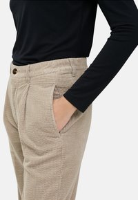 Beige corduroy pants with a ribbed texture, featuring side pockets and a button closure. Worn with a black long-sleeve top.