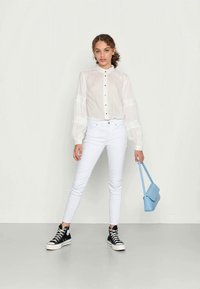 Blusa branca transparente com fecho de botão, mangas ligeiramente bufantes, combinada com jeans brancos justos e ténis de cano alto pretos. Bolsa transversal azul.