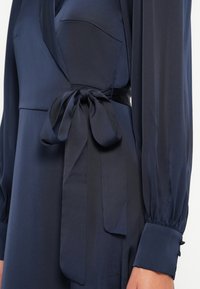 Navy wrap dress with long sleeves, featuring a waist tie, soft fabric, and a side pocket. Elegant, flowing silhouette with buttoned cuffs.