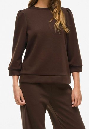 Femme portant un haut marron foncé à manches longues avec des épaules bouffantes et un pantalon assorti, debout devant un fond uni.