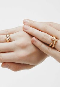 Dos manos muestran anillos: uno es de oro brillante con una piedra ovalada clara, el otro es una banda de oro retorcida con acentos en forma de pera.