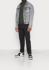 Uomo che indossa un giubbotto di jeans grigio chiaro su un dolcevita verde oliva, pantaloni neri e sneakers bianco e nere, in piedi davanti a uno sfondo bianco.
