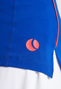 Blå atletisk topp med rosa logga som accent. Har platta sömmar och en böjd nederdel, tillverkad av stretchigt material för flexibilitet.