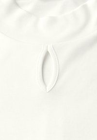Top in cotone bianco con scollo ripiegato e un piccolo dettaglio a forma di ovale sul davanti. Texture liscia e design semplice.