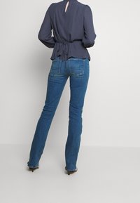 Marinebluse mit Schlüssel-Ausschnitt hinten und Schleifenakzent, kombiniert mit eng anliegenden blauen Jeans und schwarz gestreiften Absatzschuhen. Glatter Stoff, taillierte Passform.