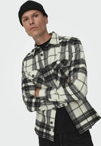 Camisa de cuadros en blanco y negro hecha de una mezcla de lana, con cierre de botones, dos bolsillos en el pecho y un corte relajado, llevada sobre una camiseta negra.