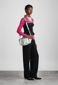 Stribet skuldertaske med et grønt, hvidt og lilla mønster; har en lang rem og er rektangulær i formen. Bæres med en sort peplum top og en pink top.