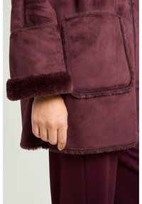 Bourgognefarvet pelsfrakke i imiteret pels med blød tekstur, brede manchetter, lappelommer og kontrasterende pelskant langs kanterne for ekstra detaljer.