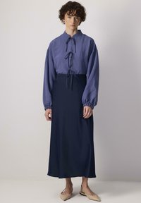 Blouse à rayures bleues avec un nœud à l'avant, manches amples ; associée à une jupe longue unie bleu foncé. Des chaussures plates beige complètent la tenue.
