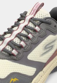 Gros plan d'une basket gris clair et crème avec des lacets gris foncé, une languette en tissu à motifs et un logo stylisé "S" sur le côté.