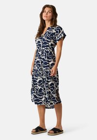 Navy Kleid mit abstraktem weißen Muster, knielang, mit kurzen Ärmeln, V-Ausschnitt, seitlichen Schlitzen, kombiniert mit schwarzen Sandalen und einem natürlich aussehenden Sohlen.