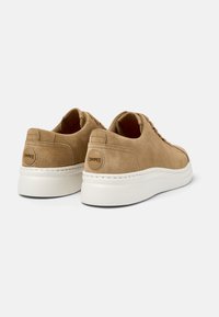 Sneakers in suede beige con suola piatta bianca, occhielli rotondi e un accento con logo circolare sul tallone. Design pulito e dettagli minimali.