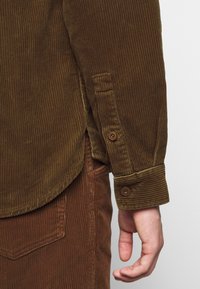 Main posée à côté d'un pantalon en velours côtelé marron et d'une manche de chemise en velours côtelé marron avec boutonnière au poignet et fente latérale.