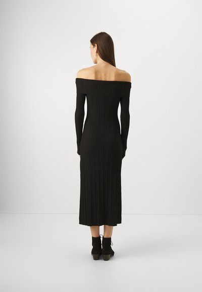 Rochie neagră, cu ștrasuri pe umeri, cu mâneci lungi, corset ajustat și lungime midi, asortată cu botine negre pe o suprafață netedă.