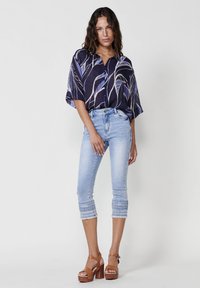 Camicia blu navy con motivi a foglia, abbinata a jeans chiari con orli ricamati, e sandali platform marroni.