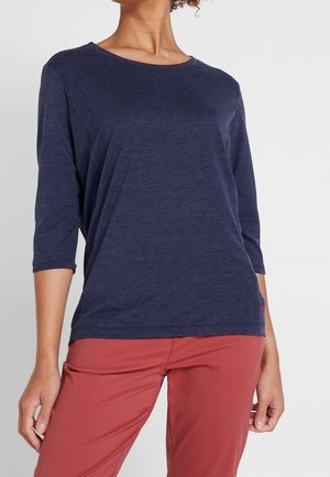 Kobieta ubrana w granatową bluzkę z rękawami do połowy przedramienia i rdzawobrązowe spodnie, stojąca na tle jednolitego jasnego tła.