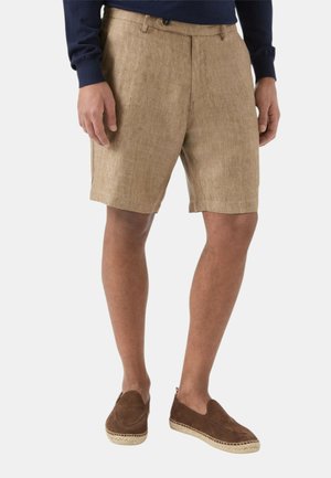 Mand iført beige linnedshorts, mørkeblå langærmet skjorte og brune slip-on espadrilles, står med afslappede hænder.