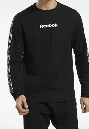 Mann trägt schwarzen gerippten Reebok-Sweatshirt mit weißem Logo auf der Brust und gebrandeten Streifen an beiden Ärmeln.