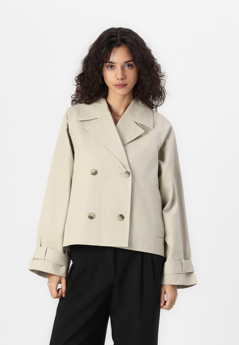 Light beige double-breasted jacket with large buttons, wide lapels, and bell-shaped cuffs, made of smooth, structured fabric.
