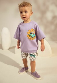 Hellviolettes übergroßes T-Shirt mit "Hot Wheels"-Grafik, kombiniert mit karierten Shorts in Weiß und Grau sowie violetten Slip-On-Schuhen.