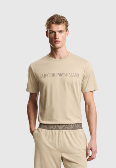 Άνδρας που φοράει μπεζ μπλουζάκι Emporio Armani και ταιριαστό σορτς με εκτύπωση λογότυπου στο στήθος και στη μέση, στέκεται μπροστά σε απλό φόντο.
