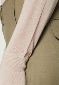 Hellrosa gestrickter Ärmel eines Pullovers, kombiniert mit einem olivgrünen Stoff. Der Pullover enthält ein dezentes Logo in einer helleren Nuance am Bündchen.