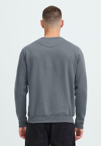 Man met kort haar, gekleed in een effen grijze lange mouwen sweatshirt en zwarte jeans, gezien van achteren tegen een lichte achtergrond.