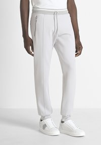 Pantalones joggers gris claro con cintura ajustable y cremalleras laterales. Hechos de material suave, con costuras en contraste. Combinados con zapatillas blancas.