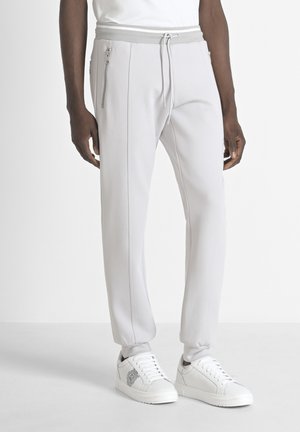 Pantalones joggers gris claro con cintura ajustable y cremalleras laterales. Hechos de material suave, con costuras en contraste. Combinados con zapatillas blancas.