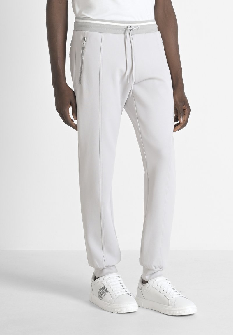 Pantalones joggers gris claro con cintura ajustable y cremalleras laterales. Hechos de material suave, con costuras en contraste. Combinados con zapatillas blancas.