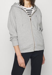 Sweat à capuche zippé gris clair en tissu doux, doté d'une capuche, de cordons de serrage et de poignets côtelés. Porté par-dessus un t-shirt à rayures noir et blanc.