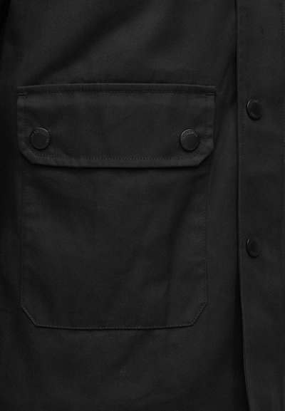 Μαύρο εξωτερικό ένδυμα με τσέπη με καπάκι και δύο μαύρα κουμπιά snap. Το υλικό φαίνεται υφή και ανθεκτικό, με καθαρό, μίνιμαλ σχεδιασμό.