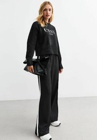 Sweatshirt corto nero e peloso con la scritta "C'EST BON", abbinato a pantaloni neri larghi con strisce bianche sui lati. Porta una borsa nera con texture.