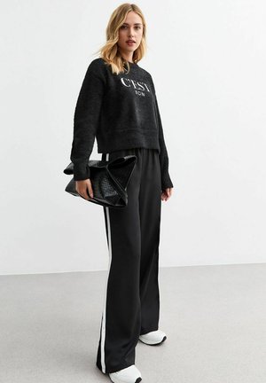 Zwarte fuzzy cropped sweatshirt met de tekst "C'EST BON", gecombineerd met losse zwarte broek met witte zijstrepen. Draagt een zwarte gestoffeerde tas.