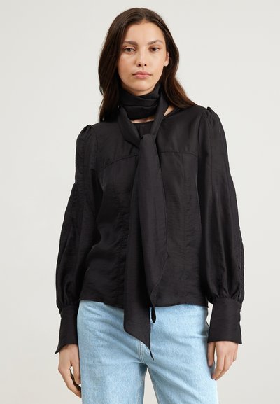 Zwarte blouse met lange mouwen van lichtgewicht stof, voorzien van een centrale strik, geplooide schouders en knoopmanchetten. Gecombineerd met lichtblauwe jeans.