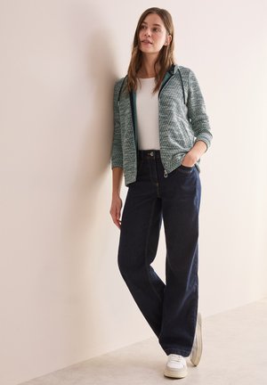 Veste zippée légère à motifs verts avec capuche, associée à un jean large bleu foncé et des baskets blanches, sur fond neutre.