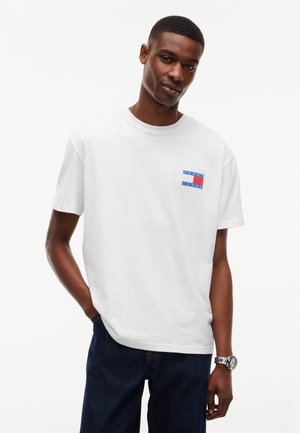 Mladý muž nosiaci biele tričko s krátkym rukávom s malým logom Tommy Jeans a tmavomodré džínsy, stojaci s jednou rukou v vrecku.