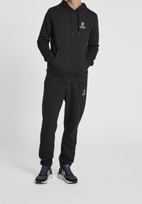 Černá mikina a kalhoty z měkké látky, které mají přední kapsu a logo na hrudi a na kalhotách. V kombinaci s černými sportovními botami.