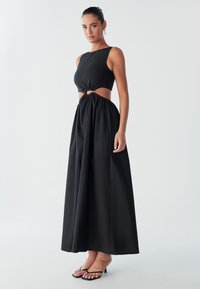 Model draagt een zwart, mouwloos maxijurkje met een uitsnede in de taille, voorzien van een geknotte detail. De achtergrond is eenvoudig en licht.