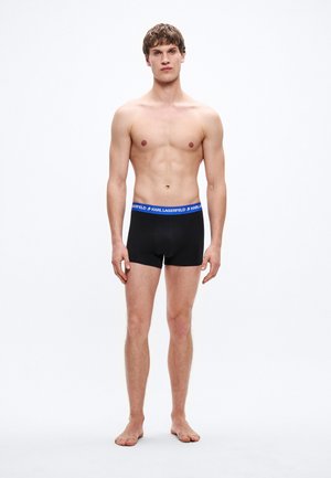 Boxer negros con una cinturilla azul que lleva el texto "KARL LAGERFELD". Tela suave, diseño ajustado, largo hasta la mitad del muslo. Modelo de pie descalzo.