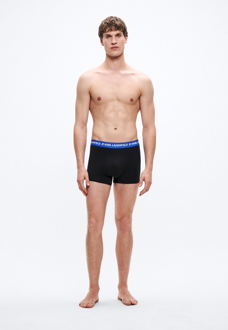 Μαύρα ανδρικά μποξεράκια με μπλε ζώνη μέσης που φέρει το κείμενο "KARL LAGERFELD". Λείο ύφασμα, εφαρμοστό σχέδιο, μήκος μέχρι τη μέση του μηρού. Το μοντέλο στέκεται ξυπόλητο.
