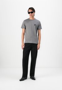 Man wearing black sunglasses, grey short-sleeve t-shirt with chest logo, black cargo pants, and black pointed-toe shoes standing against white background.