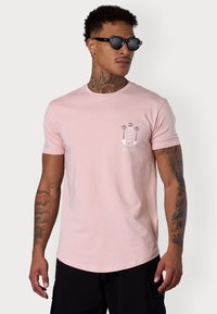 Light pink cotton t-shirt with short sleeves, crew neck, and a small white palm tree graphic on the left chest. Black cargo shorts and sunglasses.