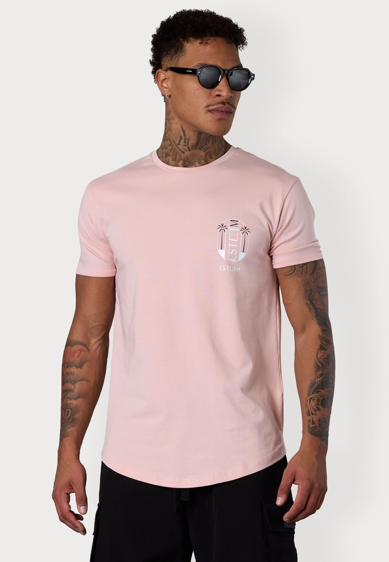 Light pink cotton t-shirt with short sleeves, crew neck, and a small white palm tree graphic on the left chest. Black cargo shorts and sunglasses.