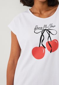 Hvid kortærmet t-shirt med røde kirsebærgrafikker og sorte bånd samt teksten "Dormir Ma Chérie"; glat bomuldsstof.