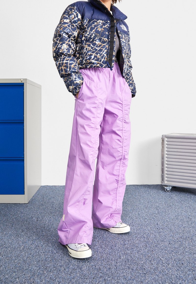 Personne portant une doudoune courte à motifs, un pantalon lavande ample et des baskets blanches, debout sur un tapis bleu près d'une armoire bleue et d'une valise argentée.