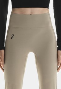 Beige High-Waist-Leggings aus dehnbarem Stoff mit einem elastischen Taillenbund und einem kleinen schwarzen Logo auf der linken Seite.