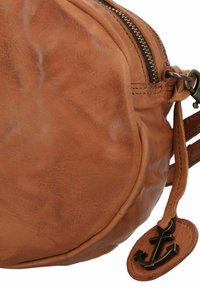 Borsa rotonda in cuoio marrone con cerniera, superficie strutturata e un'etichetta a forma di ancora appesa. Mostra dettagli di cucitura e imperfezioni naturali.