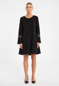 Schwarzes knielanges Kleid mit langen Ärmeln, das Lederakzente an den Manschetten, einen runden Ausschnitt und eine ausgestellte Silhouette aufweist.