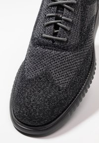 Scarpa sportiva nera con tomaia in maglia traspirante e testurizzata, punta arrotondata e suola in gomma flessibile. Chiusura con lacci prominente.